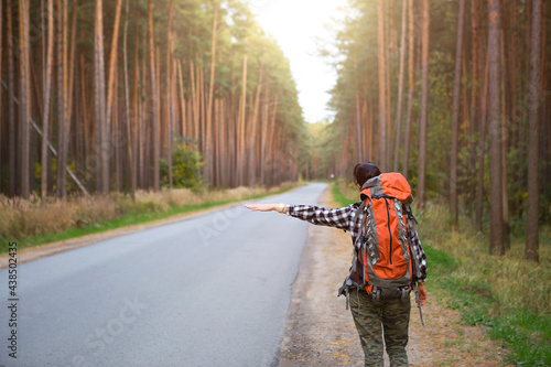 Female tourist in a checked shirt with an orange large backpack near a highway in the woods votes to get a ride. Hitchhiking, domestic tourism. Backpacker, adventure alone, trip, hike