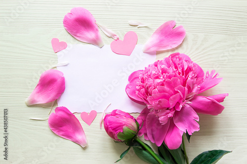 Fototapeta Naklejka Na Ścianę i Meble -  on a light wooden background there is a white card with a place for text, and next to it there are flowers of pink peonies, hearts, rose and peony petals. copy space