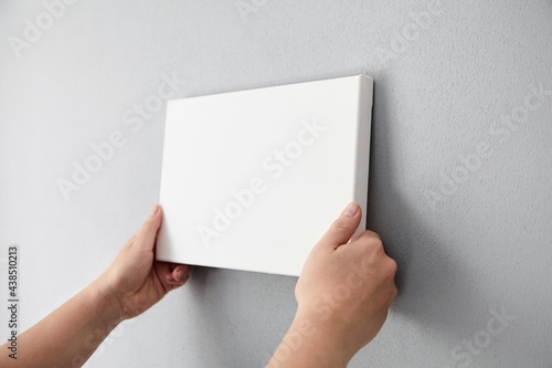 White canvas in female hands with gray wall background. Woman hanging blank picture mockup on wall