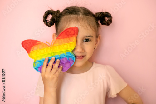 A beautiful cute little girl on a pink background covers one eye with a popit toy in the shape of a butterfly. Funny pigtails for a girl . Children's entertainment and educational toys. simple dimple photo