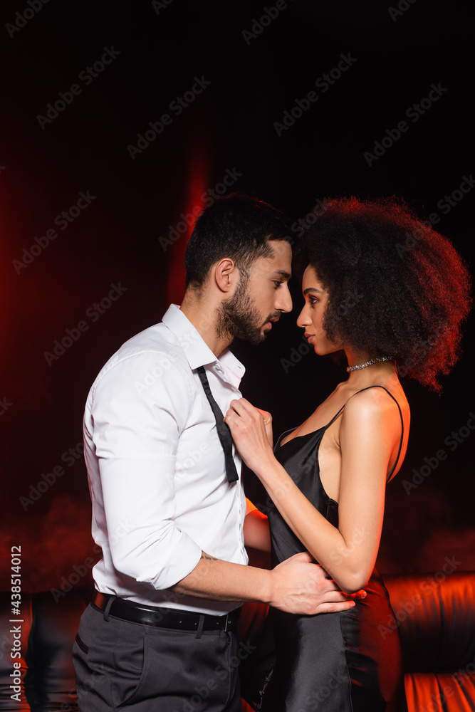 Fototapeta premium Side view of man hugging african american girlfriend in dress on black background with red lighting