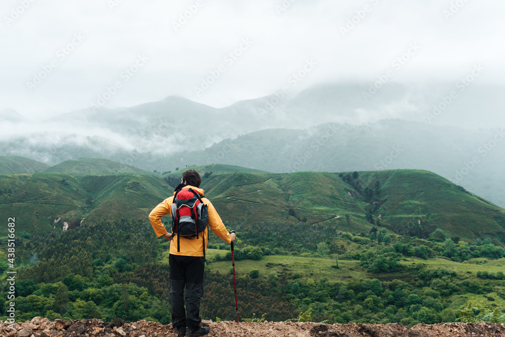 Hiker with yellow raincoat leaning on his trekking pole resting and contemplating the mountainous landscape with fog on a rainy day in Asturias, Spain. Mountain Sports