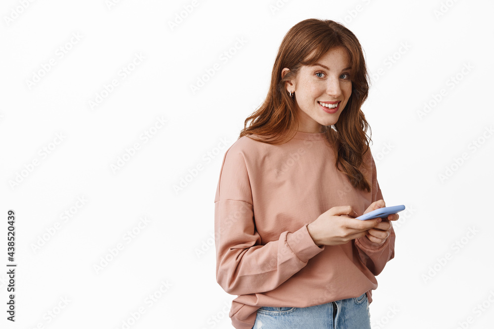 Fototapeta premium Image of smiling teenage girl with red hair, chatting on phone, messaging with smartphone and looking friendly at camera, standing over white background