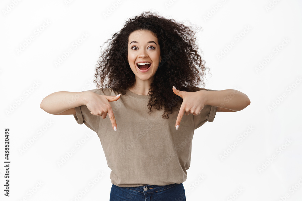 Excited beautiful woman showing announcement, pointing fingers down at advertisement and smiling amazed, happy to announce discounts in store, standing against white background Excited beautiful woman showing announcement, pointing fingers down at advertisement and smiling amazed, happy to announce discounts in store, standing against white background