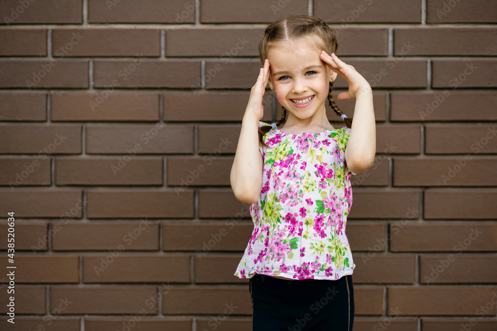 Beautiful cheerful little girl child smiles and holds her hands near her head. A little girl in summer bright clothes is standing against a brick wall and smiling cute. Free space for text