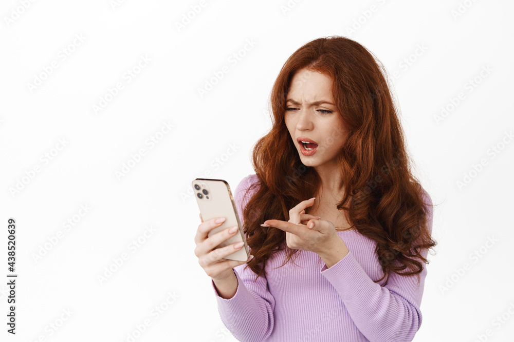 Portrait of redhead woman pointing at mobile screen with confused face, found something strange on smartphone, cant understand application, standing against white background Portrait of redhead woman pointing at mobile screen with confused face, found something strange on smartphone, cant understand application, standing against white background