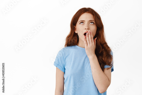 Tired and annoyed redhead girl, yawning and roll eyes uninterested, standing sleepy and unamused, hear boring story, standing against white background