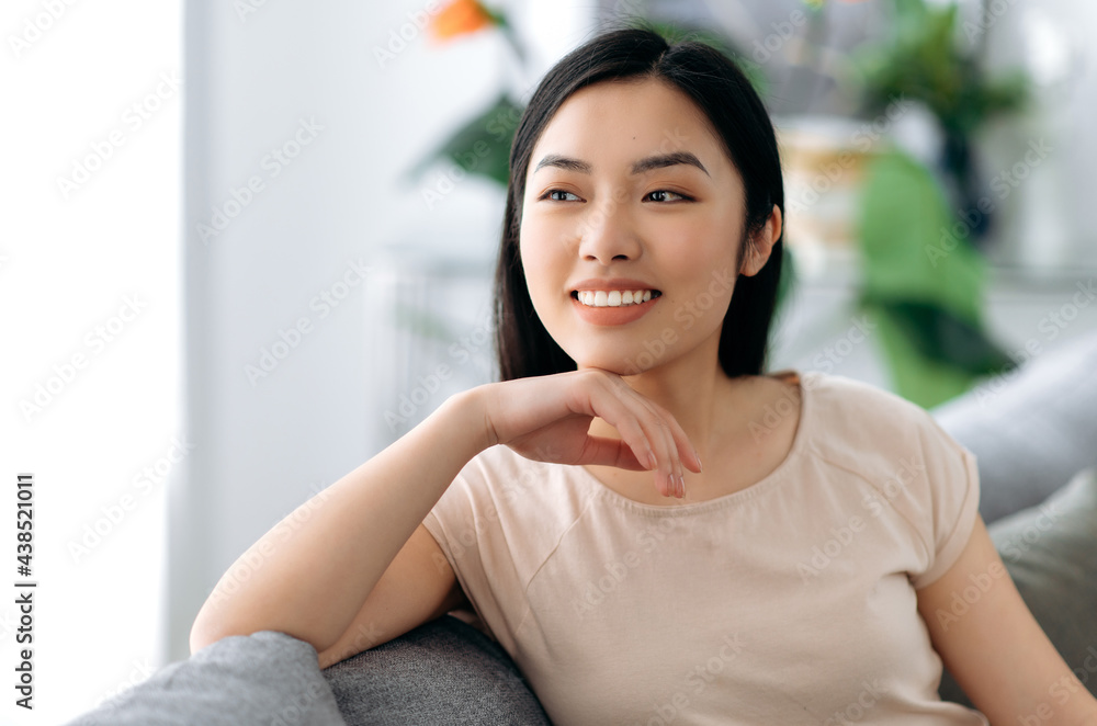 Portrait of a pretty, healthy, positive asian brunette girl, in casual t-shirt, sitting on a sofa at home, relaxing, looking away, dreaming about vacation, smiling happily Portrait of a pretty, healthy, positive asian brunette girl, in casual t-shirt, sitting on a sofa at home, relaxing, looking away, dreaming about vacation, smiling happily