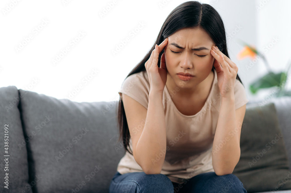 Sad asian brunette woman in stress, sitting on the sofa alone, in casual clothes, experiencing headache, migraine, massaging her temples, closing her eyes, need rest and treatment Sad asian brunette woman in stress, sitting on the sofa alone, in casual clothes, experiencing headache, migraine, massaging her temples, closing her eyes, need rest and treatment