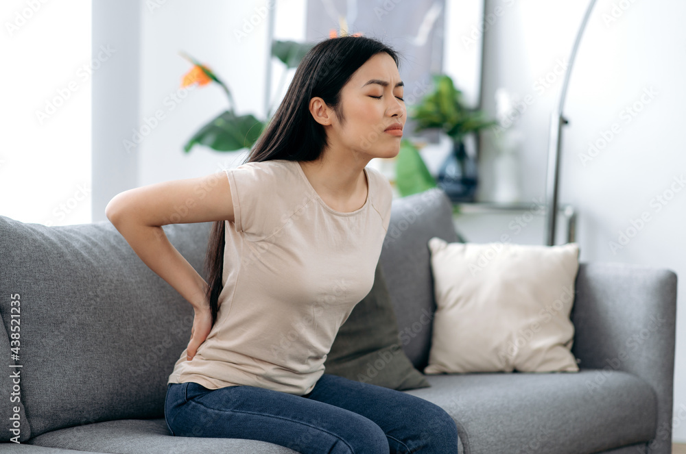 Upset frustrated asian young woman suffering from backache, unhappy japanese brunette sitting on a sofa in living room, feeling discomfort because of pain in back, need treatment and rest Upset frustrated asian young woman suffering from backache, unhappy japanese brunette sitting on a sofa in living room, feeling discomfort because of pain in back, need treatment and rest