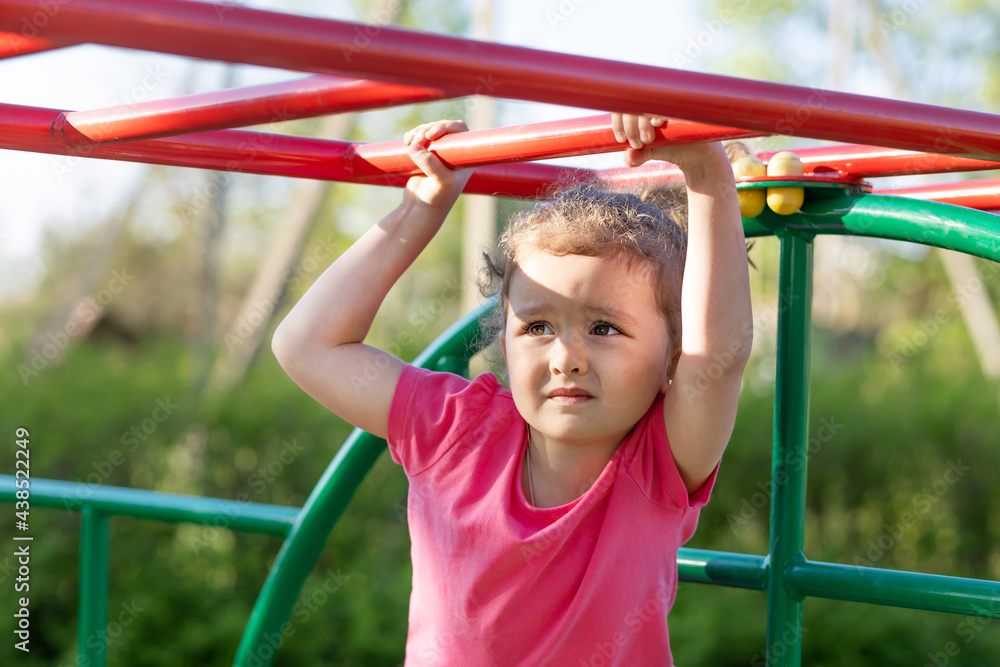 child and sport. little active emotional pensive girl on playground in park, hanging on horizontal bar, doing sports exercise outdoors. happy childhood. Kid play on school kindergarten yard in summer child and sport. little active emotional pensive girl on playground in park, hanging on horizontal bar, doing sports exercise outdoors. happy childhood. Kid play on school kindergarten yard in summer