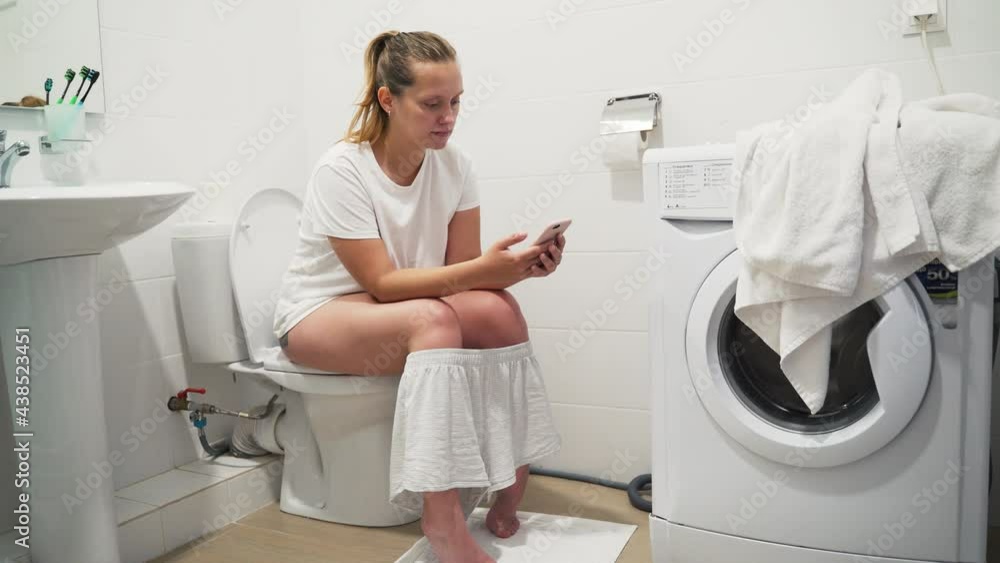 Happy beautiful blonde woman in pajamas smiles and holds smartphone in hands, sitting on the toilet. Chatting and gaming on the gadget. Lifestyle with phone. Using smartphone in everyday life Happy beautiful blonde woman in pajamas smiles and holds smartphone in hands, sitting on the toilet. Chatting and gaming on the gadget. Lifestyle with phone. Using smartphone in everyday life