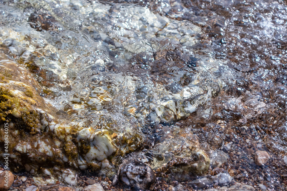 Obraz premium Wet marble stones close-up on wild mediterranean sea pebble beach. Tide on rocky shore in clear water with sun beam. Travel Greece near Athens. Natural macro patterns