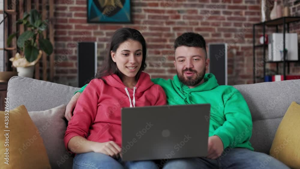Young happy caucasian couple sitting on couch and talking with family or friends online using web camera on laptop. Remote communication. Carefree lifestyle. Couple teenagers in video call conference. Young happy caucasian couple sitting on couch and talking with family or friends online using web camera on laptop. Remote communication. Carefree lifestyle. Couple teenagers in video call conference.