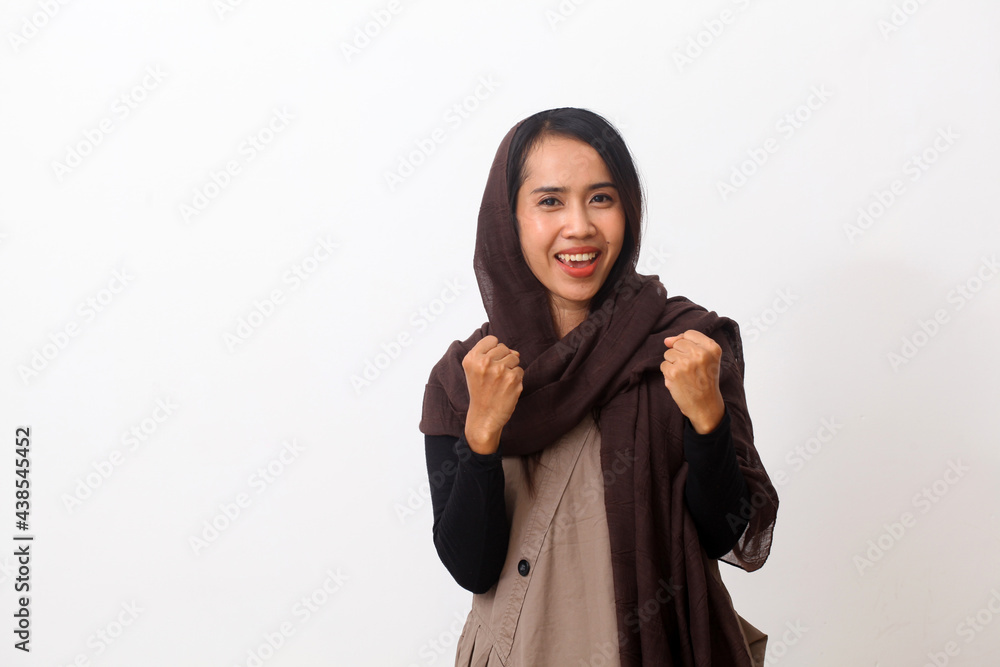 A portrait of happy asian muslim woman wearing a brown veil or hijab and white dress raises clenched fists, rejoices victory, satisfied with good result of work. White background with copy space A portrait of happy asian muslim woman wearing a brown veil or hijab and white dress raises clenched fists, rejoices victory, satisfied with good result of work. White background with copy space
