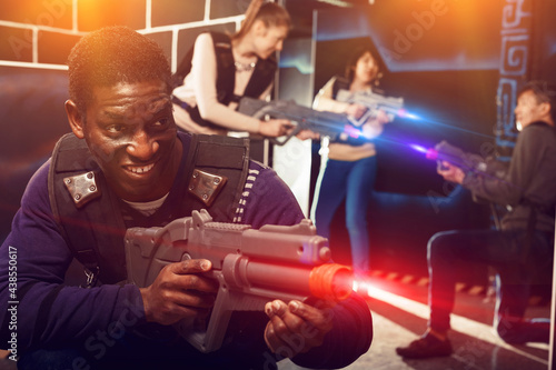 Emotional African American man aiming laser pistol at other players during lasertag game in dark room