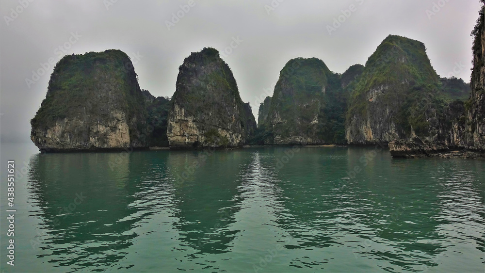 Islands with bizarre outlines hide in the fog. Green vegetation on the steep slopes. Reflection on the water. Halong Bay. Vietnam Islands with bizarre outlines hide in the fog. Green vegetation on the steep slopes. Reflection on the water. Halong Bay. Vietnam