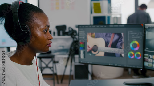 Beautiful african woman videographer with headphones editing video footage on pc with two displays working in creative agency. Worker using post production software working in creative agency office.