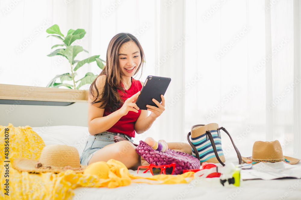 Happy Asian woman playing tablet with many items and prepare to traveling beach trip on holiday. Young cute woman using tablet to searching for a trip on weekend at home.