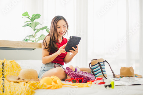 Happy Asian woman playing tablet with many items and prepare to traveling beach trip on holiday. Young cute woman using tablet to searching for a trip on weekend at home.