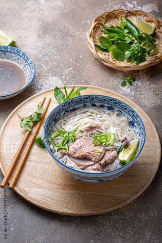 Traditional vietnamese noodle soup pho in bowl, garnished with basil, mint, lime, on concrete background