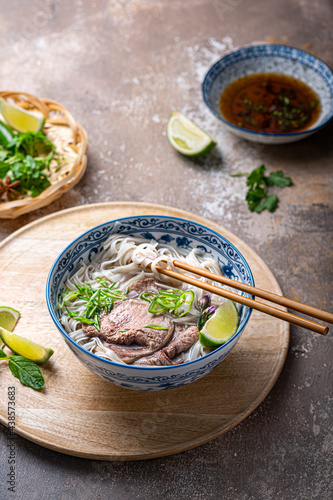 Traditional vietnamese noodle soup pho in bowl, garnished with basil, mint, lime, on concrete background