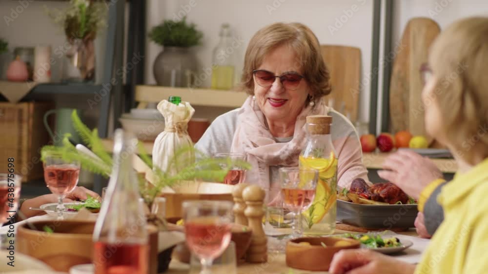 Positive elderly women sitting at dinner table, enjoying meal and speaking with friends at home party Positive elderly women sitting at dinner table, enjoying meal and speaking with friends at home party