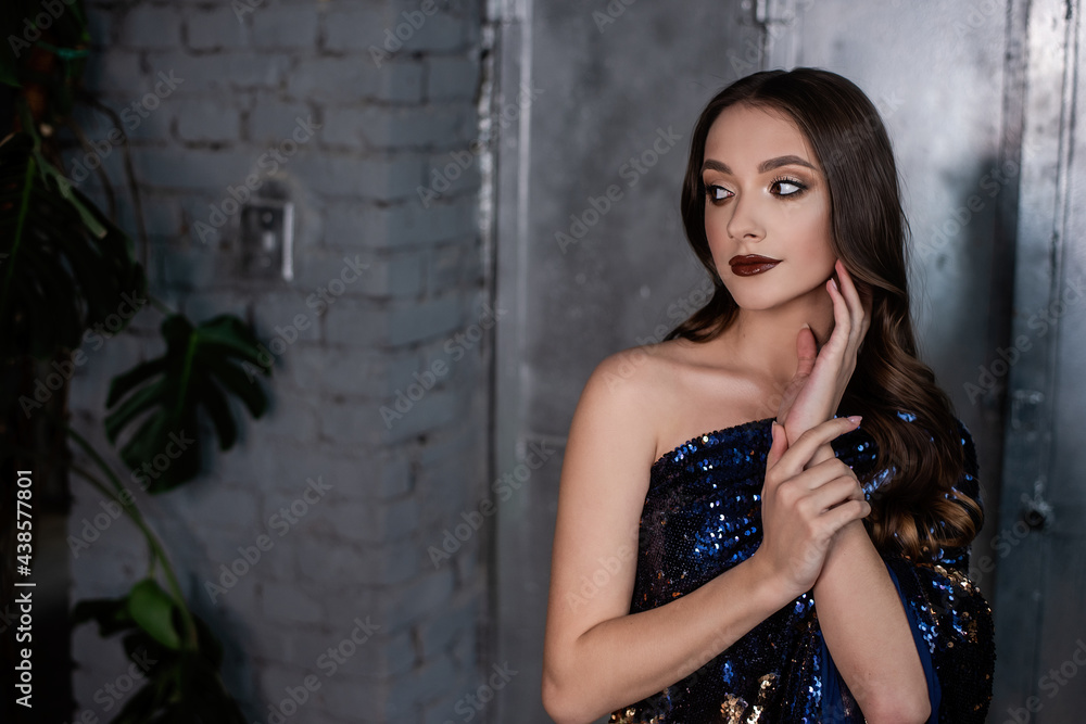 Fototapeta premium Close-up portrait of beautiful girl with an evening festive professional make-up in blue, gold dress in sequins on metallic, gray shiny background. Grunge texture of interior. Clean skin, healthy face