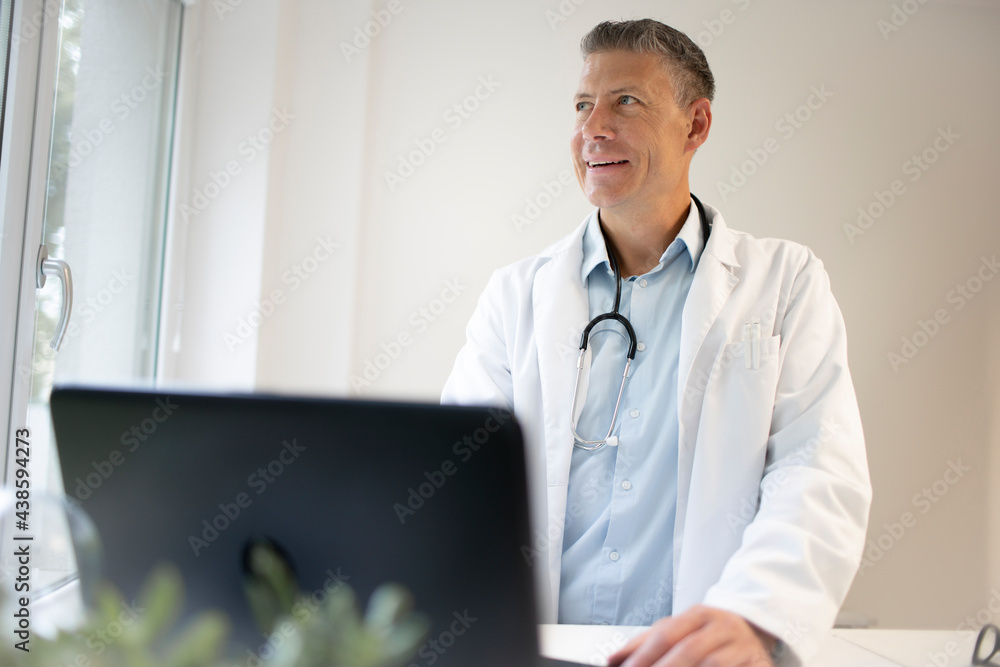 handsome doctor with white coat and blue shirt and stethoscope stands behind high table and works on laptop and has web conference or meeting