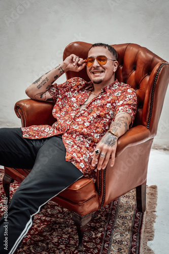 Expressive and stylish guy in hawaiian shirt and sunglasses relaxing on a luxury retro sofa looks like freakly gangster