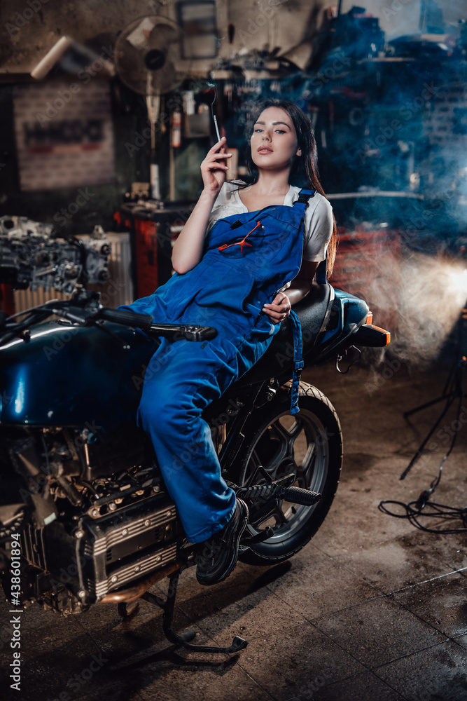 Female mechanic relaxing smoking a cigarette while lying on custom bobber in garage or workshop Female mechanic relaxing smoking a cigarette while lying on custom bobber in garage or workshop