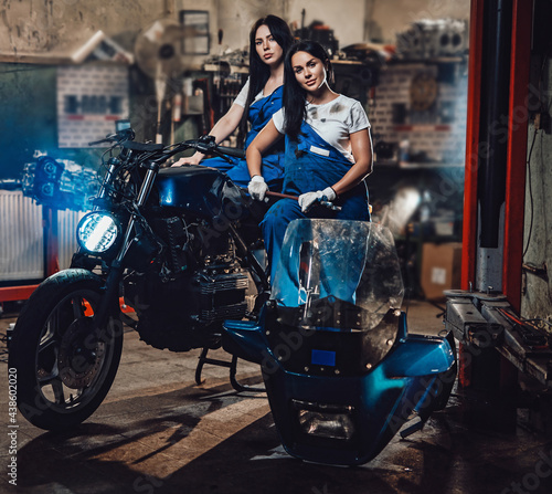 Two brunette women in blue overalls posing next to a custom bobber in authentic workshop garage
