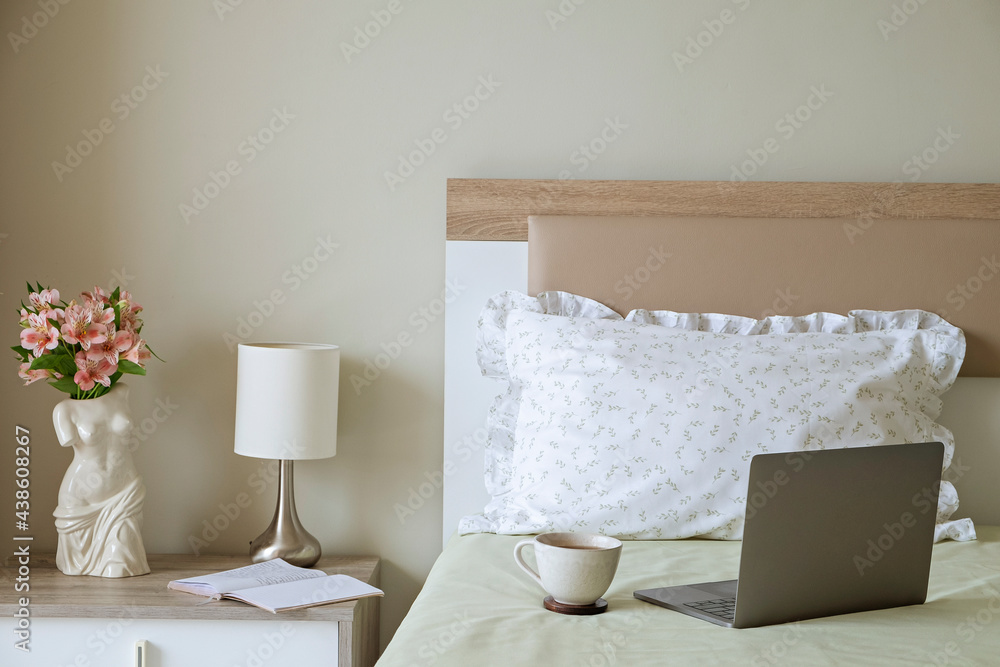 Good morning concept. Empty unmade bed with laptop with open lid, cup of coffee and alstroemeria flowers in venus de milo sculpture shaped vase on a nightstand. Close up, copy space, background. - obrazy, fototapety, plakaty 