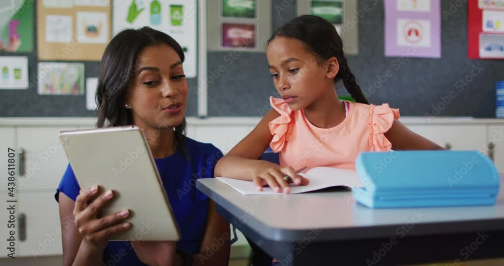 Diverse female teacher and schoolgirl in classroom, looking at tablet, smiling Diverse female teacher and schoolgirl in classroom, looking at tablet, smiling