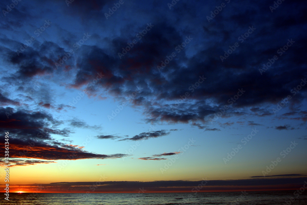 The twilight blue-scarlet sky at sunset with clouds, which turned from light to black, over the calm sea, the edge of the setting sun can be seen on the horizon.
