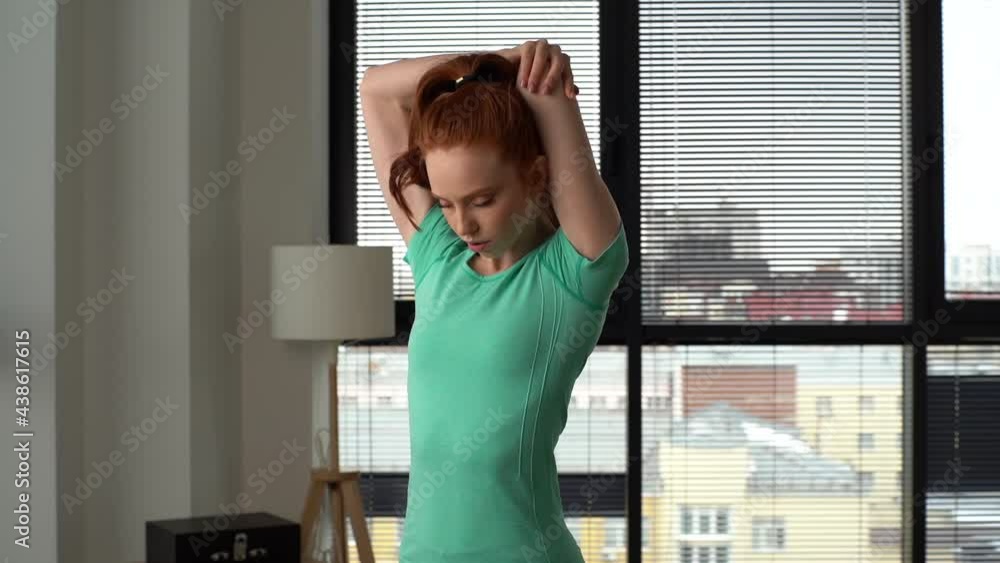 Tracking shot of sporty young woman doing stretching exercise before fitness training at home office on background of window in sunny day. Fitness redhead female warming up before muscles in apartment Tracking shot of sporty young woman doing stretching exercise before fitness training at home office on background of window in sunny day. Fitness redhead female warming up before muscles in apartment