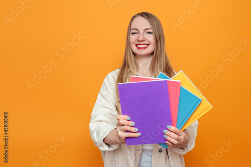 Caucasian blonde student girl holds four books in multi-colored covers smiling isolated over orange background in studio. English language school, education concept