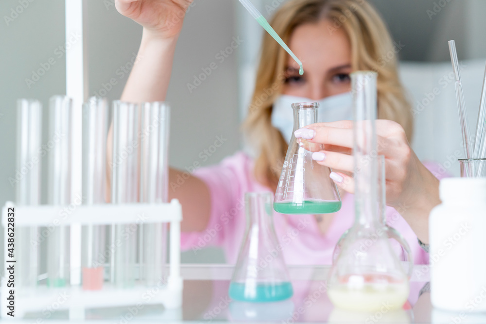 female microbiologist in pink coat conducts research in laboratory, drops green liquid with pipette into flask with colored liquid, blured foreground. female microbiologist in pink coat conducts research in laboratory, drops green liquid with pipette into flask with colored liquid, blured foreground.