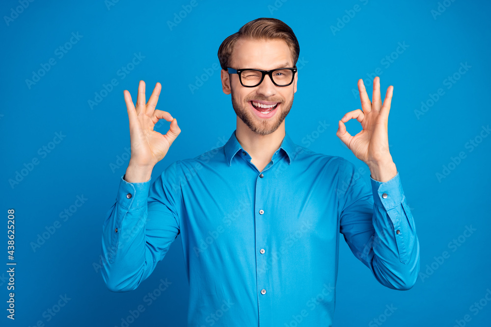 Photo of young man happy positive smile blink eye show okay alright great decision advert isolated over blue color background Photo of young man happy positive smile blink eye show okay alright great decision advert isolated over blue color background