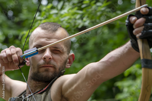 Portrait of a man aiming an arrow from a bow in the forest, close-up, selective focus. Concept: men's hobbies, survival in difficult conditions, hunting and recreation.