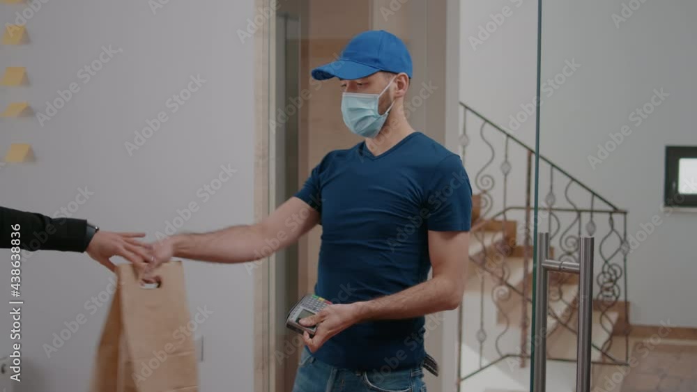 Delivery guy with protection face mask and gloves delivering takeaway food box during lunchtime in company office. Businesswoman client paying meal order with smart watch using POS contactless Delivery guy with protection face mask and gloves delivering takeaway food box during lunchtime in company office. Businesswoman client paying meal order with smart watch using POS contactless