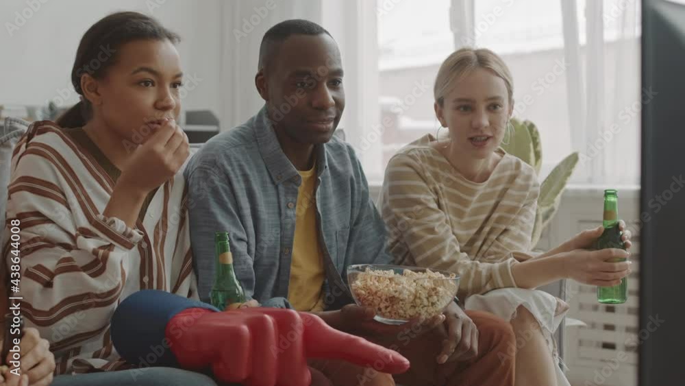 Backlighted of African man, Mixed-Race and Caucasian woman sitting on couch in living room, drinking beer and eating snacks, watching favourite sports on television Backlighted of African man, Mixed-Race and Caucasian woman sitting on couch in living room, drinking beer and eating snacks, watching favourite sports on television