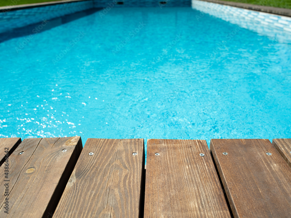 Ripped wavy water surface in swimming pool with wooden, old deck and green grass around. Swimming pool area in hotel or villa. Summer vacation background.