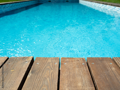 Ripped wavy water surface in swimming pool with wooden, old deck and green grass around. Swimming pool area in hotel or villa. Summer vacation background.