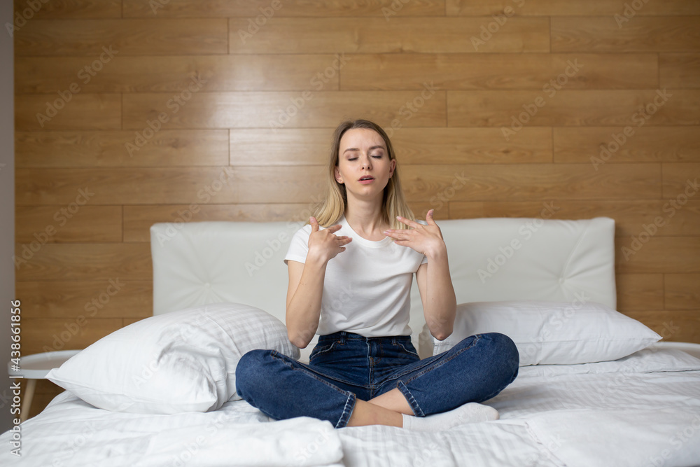 A young woman is sitting on a sofa or bed and waving her arms from the heat at home. Female suffers from stuffy and hot indoors due to lack of air conditioning or ventilation in House apartment.

