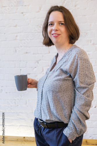 Half-length portrait of a young brunette woman with a cup of coffee in her hand against a background of a white brick wall. The woman smiles and holds a ceramic gray mug in her hand. photo