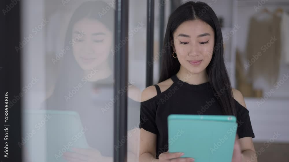 Portrait of smiling satisfied young Asian woman checking e-mail on tablet in the morning in home office with wardrobe at background. Confident positive employee on remote working indoors Portrait of smiling satisfied young Asian woman checking e-mail on tablet in the morning in home office with wardrobe at background. Confident positive employee on remote working indoors