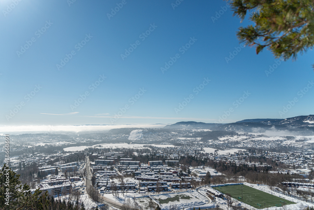 Panoramic view of a small city with mountains and clouds in the distance and a soccer football field framed by evergreen branches Panoramic view of a small city with mountains and clouds in the distance and a soccer football field framed by evergreen branches