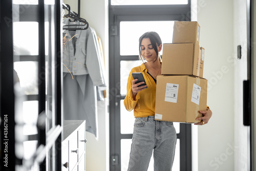 Young woman with parcels at the hallway at home, holds smart phone and checking goods. Concept of buying online and delivering goods home photo