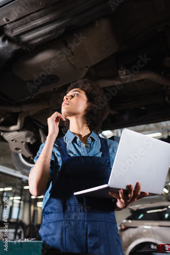 young african american mechanic inspecting lifted car with laptop in hand in garage